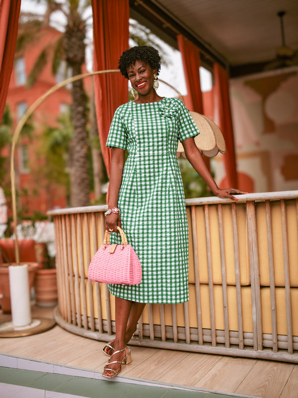 Woman in a green checkered dress holding a pink handbag on a wooden deck.