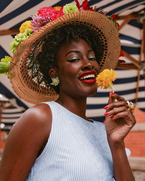 Woman wearing a straw hat with colorful flowers, holding a yellow flower, against a striped background.